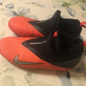 Röda/oranga Phantom fotbollsskor från Nike - Säljer ett par röda/oranga Phantom Nike fotbollsskor med svarta detaljer och en stilren design. Skorna har en strömlinjeformad form med dobbar för optimalt grepp på planen. Perfekta för fotbollsspelare som vill ha både stil och funktion. Snöre ingår men går även att använda utan snöre!