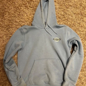 Blå hoodie från Dickies - Säljer en snygg blå hoodie från Dickies med deras logga tryckt på bröstet och ryggen. Tröjan har en klassisk känguruficka och en justerbar huva. Perfekt för en avslappnad stil.