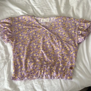 Lila blommig omlottopp från Urban Outfitters - Säljer en söt lila omlottopp från Urban Outfitters med gula blommor. Toppen har korta ärmar och en v-ringad design. Perfekt för vår och sommar! 🌸