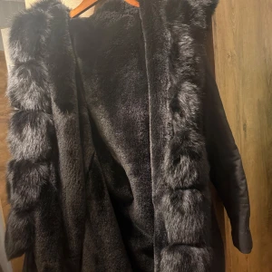 Svart pälsjacka med huva - Säljer en elegant svart pälsjacka med stor huva. Jackan har en lyxig känsla med mjuk pälsdetalj längs kanterna och runt huvan. Perfekt för att hålla sig varm och stilren under kalla dagar. Original pris: ≈ 5500kr PS: Aldrig använd!!