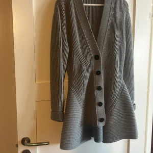 Grå stickad kofta från Alexander McQueen - Säljer en elegant grå stickad kofta från Alexander McQueen, tillverkad i Italien. Koftan har en v-ringad design med knappar framtill och långa ärmar. Den har en unik ribbad struktur som ger en stilren look.