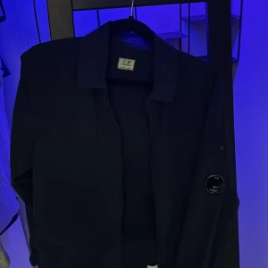 Mörkblå overshirt från CP Company - Säljer en stilren mörkblå overshirt från CP Company. Jackan har en dragkedja framtill och en praktisk ficka på bröstet. Ärmen har en unik detalj med en lins. Perfekt för en avslappnad look äkta köpt från nk, ny pris 2800 säljer den biligt för de inte är min still och inte har andvänt den