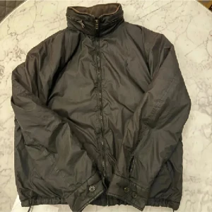 Svart vindjacka med dragkedja - Säljer en stilren svart vindjacka med dragkedja och hög krage. Jackan har långa ärmar och justerbara muddar med knappar. Perfekt för blåsiga dagar. Tappat en knapp på ena fickan 