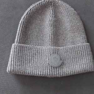 Grå ribbad mössa från Moncler - Snygg grå ribbad mössa från Moncler med uppvikt kant och logotyp framtill. Endast använt 4 gånger. Perfekt för att hålla värmen under kyliga dagar. Stilren design som passar till en "grisch" outfit. Även öppen för byten!! Tveka ej höra av dig om du har några funderingar! Skickar endast spårbart!