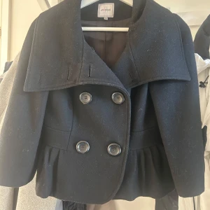 Svart kappa från Monnari - Elegant svart kappa från Monnari med stora knappar framtill och volangdetalj vid nederkanten. Jackan har en unik design med en bred krage och trekvartsärmar. Perfekt för en stilfull look.