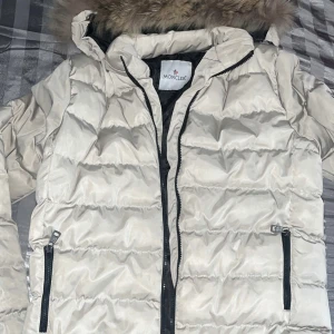 Beige dunjacka från Moncler - Säljer en snygg beige dunjacka från Moncler med pälskantad huva. Jackan har dragkedja och två fickor med dragkedjor framtill. Perfekt för kyliga dagar!