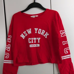 Röd croppad tröja med tryck - Säljer en röd croppad tröja från H&M med texten 'New York City' och 'Crew 94' på framsidan. Tröjan har långa ärmar med tryck och vita ränder. Perfekt för en avslappnad stil.