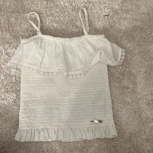 Vit topp med volang från Guess - Säljer en söt vit topp från Guess med volangdetaljer. Toppen har smala axelband och en elastisk design som ger en bekväm passform. Perfekt för varma sommardagar! 🌸 (inga defekter) ganska kort i modellen🩷