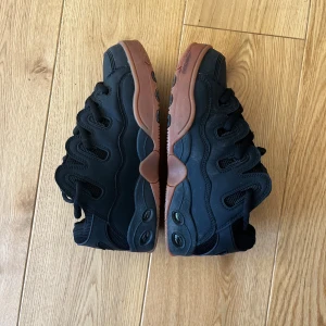 Osiris skor Svarta sneakers med brun sula - Snygga svarta sneakers med en kontrasterande brun sula. Skorna har en unik design med snörning och detaljer på sidorna. Perfekta för en cool och avslappnad stil.