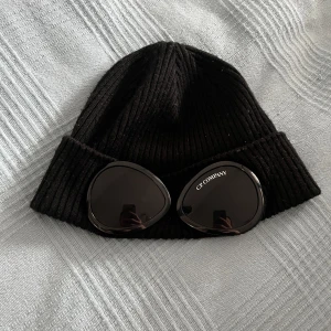 Svart ribbad mössa från C.P. Company - Använd cs 4-5 gånger! Snygg svart ribbad mössa från C.P. Company med unika glasögondetaljer framtill. Perfekt för att ge din outfit en cool touch. Tillverkad i ett bekvämt material som håller dig varm.