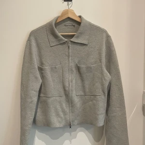 Grå stickad jacka med dragkedja från Åhléns - Säljer en stilren grå stickad jacka från Åhléns med dragkedja framtill och två stora fickor. Jackan har klassisk krage och lång ärm, perfekt för lager-på-lager. Enkel och snygg design som passar till mycket. Något sliten på dragkedjan (bild finns), annars i gott som nyskick. Nypris 500:-, säljer för 200:-
