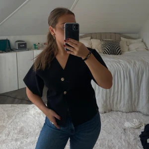 Svart blus med guldknappar - Stilren svart blus med v-ringning och korta ärmar. Blusen har två dekorativa guldknappar framtill och en rak passform. Perfekt att matcha med jeans eller kjol för en snygg look.