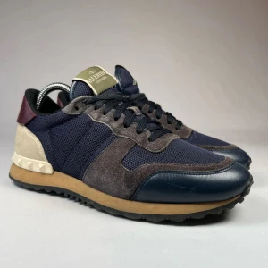 Valentino Rockrunner sneakers i blå och beige - Snygga Valentino Rockrunner sneakers med ovandel i blå mesh, mörkgrå mockadetaljer och beige häl med nitar. Skorna har svarta snören, bruna och svarta detaljer samt platt sula. Perfekta för dig som gillar exklusiva och sportiga sneakers.