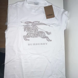 Burberry T-shirt passar som S - ”Passar som S” Säljer en vit t-shirt från Burberry med stort klassiskt ryttare-tryck och logga på bröstet. T-shirten har rund halsringning och är kortärmad. 