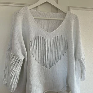 Vit stickad tröja med hjärtmönster - Säljer en vit stickad tröja med v-ringning och ett stort hjärtmönster framtill. Tröjan har en lös, oversized passform. Perfekt för dig som gillar mysiga och trendiga plagg.