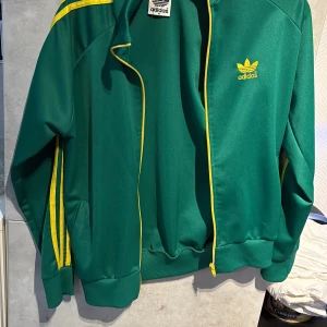 Grön Adidas track jacket med gula detaljer - Säljer en grön Adidas retro track jacket med gula ränder längs ärmarna och gul dragkedja. Jackan har broderad Adidas-logga på bröstet och klassisk ribbad mudd vid ärmslut och midja. Perfekt för en sportig stil. 