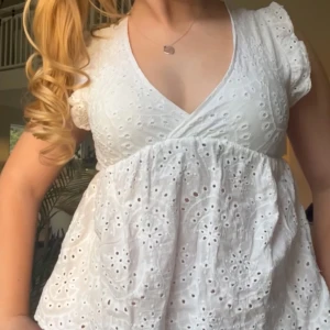 Vit omlott topp med broderade detaljer - Söt vit topp med omlott och v-ringning. Toppen har broderade spetsdetaljer och små puffärmar. Perfekt för sommarens alla tillfällen.