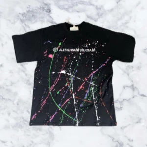 Svart t-shirt från Maison Margiela med färgstänk - Cool svart t-shirt från Maison Margiela med färgstänk i grönt, rosa, vitt, lila och orange. Tryckt logga framtill. Perfekt för dig som gillar streetwear och vill sticka ut med en unik design.