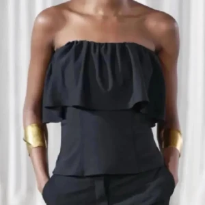 Svart axelbandslös tubtopp med volang - Säljer en svart axelbandslös tubtopp med volangdetalj upptill. Toppen har en stilren och elegant design som passar perfekt till många olika outfits. Toppen kommer från zara i nyskick.