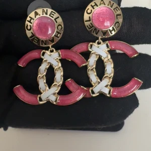 Rosa och guldiga örhängen från C - Säljer ett par statement-örhängen från C i guld med rosa detaljer. Örhängena har den klassiska C-loggan med kedjedetaljer och vita inslag. Perfekta för att lyfta vilken outfit som helst!