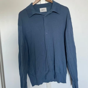 Blå långärmad pikétröja från NN07 - Säljer en stilren blå långärmad pikétröja från NN07 med klassisk krage och knäppning framtill. Tröjan har en mjuk känsla och ribbade muddar vid ärmslut och nederkant. Perfekt för en avslappnad men snygg look.