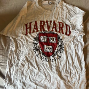 Vit Harvard t-shirt från FB Sister/ New yorker - Vit t-shirt från FB Sister/ New yorker med stort rött Harvard-tryck och emblem på bröstet. Klassisk rund hals och korta ärmar. Perfekt för dig som gillar college-stil och vill ha en enkel men snygg statement-tisha. Storlek M