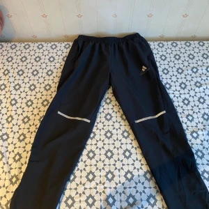 Svarta träningsbyxor från Adidas - Svarta träningsbyxor från Adidas med resår i midjan och reflexdetaljer på benen. Perfekta för löpning eller gym. Lätta och smidiga med diskret logga på låret.