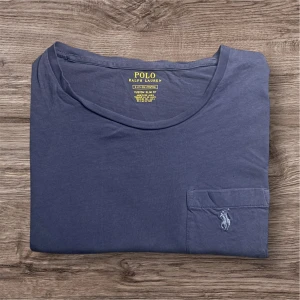 Ralph Lauren T-shirt – Blå (S)  - Ralph Lauren T-shirt – Blå (Passar ca 170 cm) 💙  Säljer en äkta Ralph Lauren t-shirt i blått med bröstficka och broderad logga – stilren och perfekt till vardags! T-shirten har några märken (syns på bilden), därav det lägre priset, men det påverkar inte användningen. 	•	👕 Storlek: Passar dig som är ca 170 cm lång 	•	🎨 Färg: Blå 	•	🧵 Detaljer: Bröstficka med broderad logga 	•	💸 Billigt pris p.g.a. märken (se bild) 	•	✅ Äkta Ralph Lauren  115