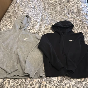Nike ziphoodies i grått och svart - Två klassiska Nike hoodies med dragkedja, en i grått och en i svart. Båda har huva, fickor framtill och Nike-logga på bröstet. Perfekta för en sportig och avslappnad stil. 100kr för en 150kr för båda