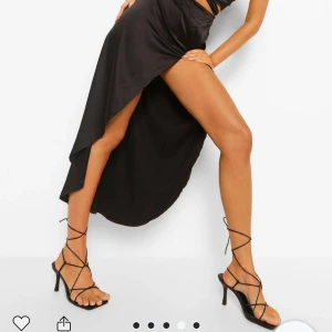 Svart asymmetrisk långklänning - Säljer en svart långklänning med asymmetrisk fåll och omlott-detalj framtill. Klänningen har hög slits och knytning i midjan för en snygg siluett. 