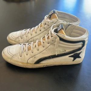 Vita höga sneakers från Golden Goose - Säljer ett par vita höga sneakers från Golden Goose med svarta detaljer. Tvärtfeta och najs färg till sommaren. Använda men med GG stil ser dom fortfarande väldigt bra ut på foten. Tyvärr inget kvitto eller box, där av lågt pris 