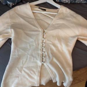 Beige kofta med knappar - Säljer en stilren beige  kofta från Gina tricot med  v-ringning och dekorativa knappar framtill. Tröjan har lång ärm och är perfekt att slänga över en topp eller klänning. Enkel och snygg design som passar till mycket.