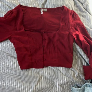 Röd blus med knappar från H&M  - Säljer en röd blus från H&M Divided med långa ärmar och knappar framtill. Blusen har en fyrkantig urringning och puffiga ärmar med knäppning vid ärmslutet. Perfekt för dig som gillar en feminin och trendig stil.
