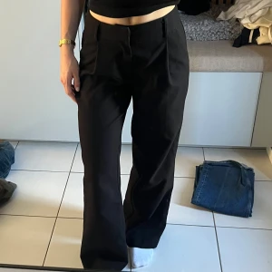 Svarta kostymbyxor med pressveck - Snygga svarta kostymbyxor med raka ben och pressveck framtill. Byxorna har hög midja och stängs med dold dragkedja och hake. Perfekta för en stilren och klassisk look.