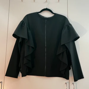 Svart blus med volangärmar - Säljer en svart blus från Vila med dragkedja framtill och unika volangdetaljer längs ärmarna. Blusen har rund halsringning och lång ärm, perfekt för dig som vill sticka ut med en stilren men ändå lekfull look. Har används ett få antal gånger men inga synliga tecken på det. Storleken är M. Priset kan diskuteras. 