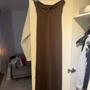 Brun axelbandslös långklänning - Säljer en brun axelbandslös långklänning med twistad detalj vid bysten. Klänningen har en elegant och enkel design med öppen rygg