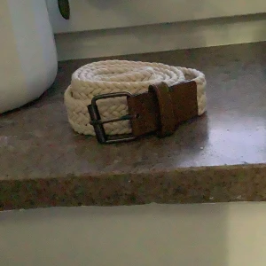 Beige flätat bälte med brunt spänne - Säljer ett beige flätat bälte med ett klassiskt metallspänne och detaljer i brunt läder. Bältet har en stilren design och passar perfekt till jeans eller chinos. Enkel att justera tack vare det flexibla materialet.