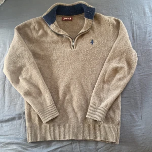 Beige stickad half zip  - Säljer en beige stickad tröja från MC med halv dragkedja och mörkblå krage. Tröjan har lång ärm, ribbade muddar och en liten broderad logga på bröstet. Perfekt för lager-på-lager under kyliga dagar. Vid ytligare fårgor är de bara att höra av sig, pris kan sänkas vid snabb affär. 