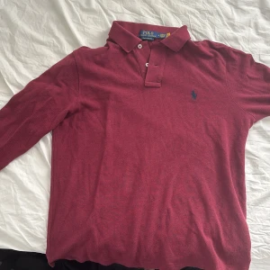 Vinröd pikétröja från Polo Ralph Lauren - Säljer en vinröd långärmad pikétröja från Polo Ralph Lauren i modellen Custom Slim Fit och är i storlek M. Tröjan har klassisk krage, knappar vid halsen och den ikoniska blå broderade loggan på bröstet.
