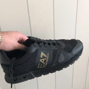 Svarta sneakers från EA7 Emporio Armani - Säljer ett par svarta sneakers från EA7 Emporio Armani med guldfärgad logga på sidan. Skorna har snörning, rund tå och platt sula. Materialet är en mix av syntet och mesh för en sportig look.