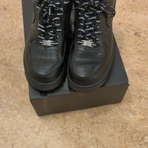 Nike Air Force 1 x Supreme svarta sneakers - Svarta Nike Air Force 1 sneakers i samarbete med Supreme. Skorna har Supreme-logga på sidan och svarta skosnören med vit Supreme-text. Klassisk rund tå och perforerad tåbox för extra ventilation. Perfekta för dig som gillar streetwear.