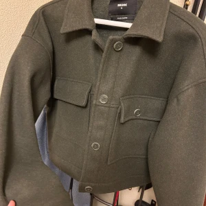 Mörkgrön ullblandad jacka från BikBok - Säljer en mörkgrön jacka från BikBok i ullblandning. Jackan har stora bröstfickor med knappar, klassisk krage och knäppning framtill. Modellen är croppad och har långa ärmar. Perfekt för lager-på-lager-stil.