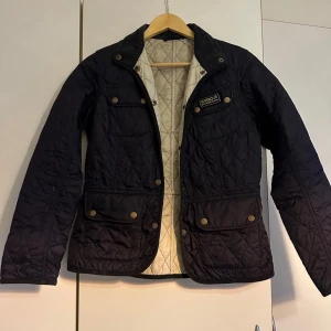 Marinblå quiltad jacka från Barbour - Säljer en marinblå quiltad jacka från Barbour med guldiga knappar och fyra fickor framtill. Jackan har lång ärm och klassisk krage, samt ljus insida med rutmönster. Perfekt för dig som vill ha en stilren och tidlös look.