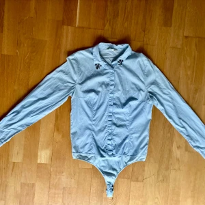 Ljusblå skjorta bodies från КФТФП i storlek 42 - En långärmade ljusblå body-skjorta med dekorativa detaljer vid kragen från КФТФП i storlek 42. Den är en mångsidigt plagg med en smickrande passform. Skjortan är gjord av bomull, nylon och elastan.