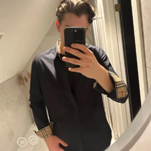 Svart skjorta från Burberry med rutiga detaljer - Säljer en svart skjorta från Burberry med klassiska rutiga detaljer på insidan av kragen och ärmsluten. Skjortan har knappar framtill och en diskret broderad logga på bröstet. Tillverkad i 100% bomull.