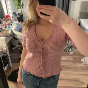 Rosa ribbad topp från ONLY - Säljer en söt rosa ribbad topp från ONLY med volangdetaljer längs kanterna och knappar framtill. Toppen är croppad och har korta ärmar samt v-ringning. Perfekt för vår och sommar!