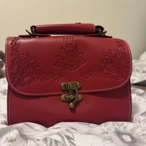 Röd handväska från Anna Sui - Säljer en snygg röd handväska från Anna Sui med dekorativa mönster på framsidan och ett unikt metallspänne. Väskan har ett handtag upptill och ett svart innerfoder med innerficka med dragkedja. Perfekt accessoar för att lyfta din outfit.