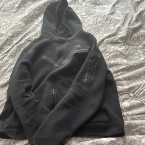 Nike tech - Säljer en svart hoodie med dragkedja framtill och huva. Tröjan har en ficka med dragkedja på ena ärmen och är tillverkad i ett mjukt material. Perfekt för kyliga dagar eller som ett extra lager.