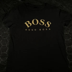 Hugo boss t-shirt - Svart t-shirt från Hugo Boss med stor guld BOSS-logga. Klassisk passform och rund halsringning. Perfekt för dig som gillar stilrena märkesplagg. Passa på o skaffa ett snyggt plagg till sommaren för bara 249 kr + frakt