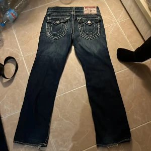 True Religion Ricky Super T jeans - Säljer ett par mörkblå True Religion Ricky Super T jeans med vit kontrastsöm och klassiska bakfickor med lock och knapp. Jeansen har tydliga detaljer på fickorna. Perfekta för dig som gillar knappt använda nyttskick jeans. 